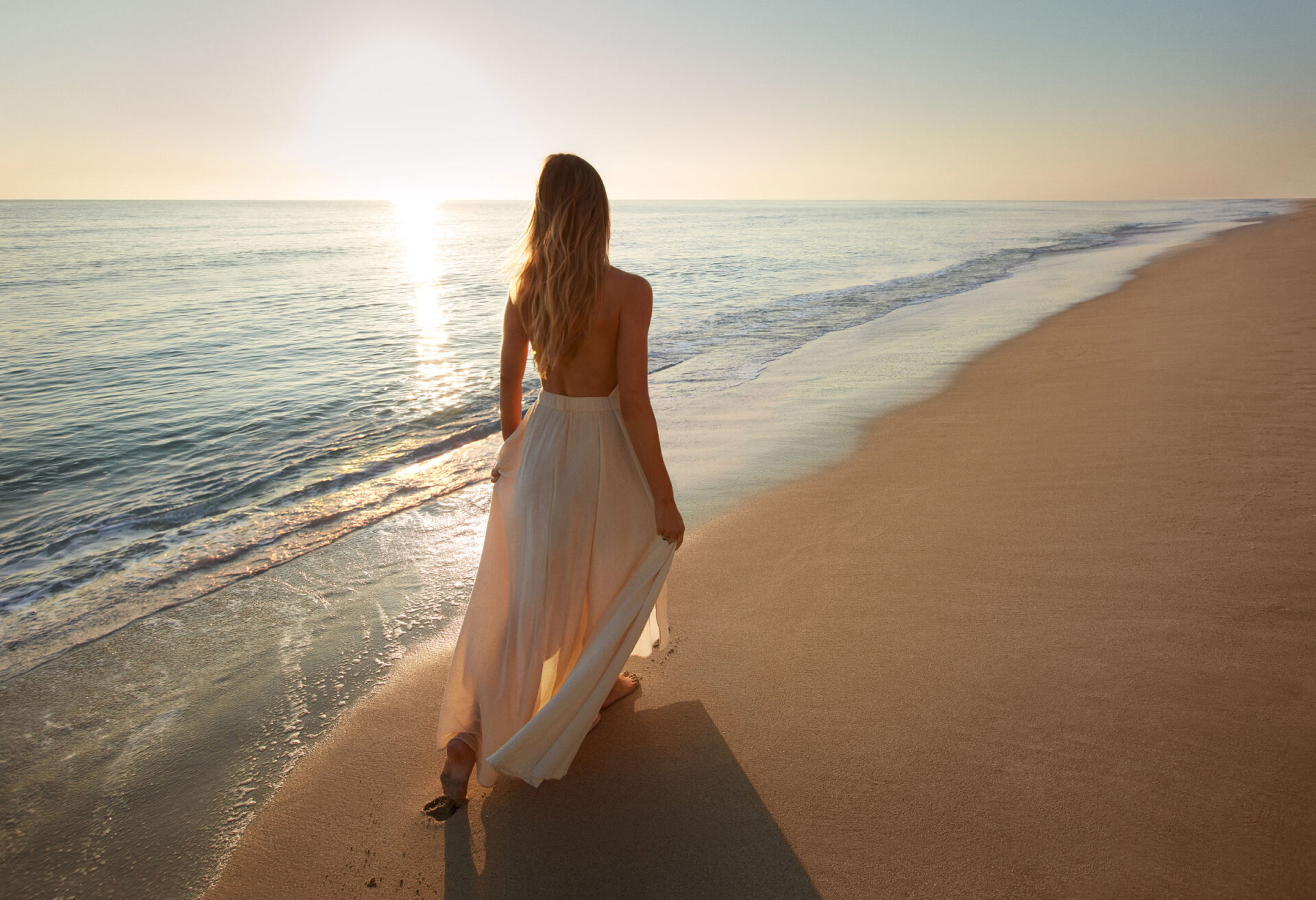 A woman in a flowing white dress walks along an empty beach at sunset, with the sun low over the horizon and gentle waves on the shore.